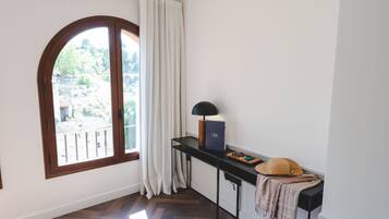 Una habitación con un amplio ventanal arqueado, un escritorio negro con lámpara, un libro y un sombrero.