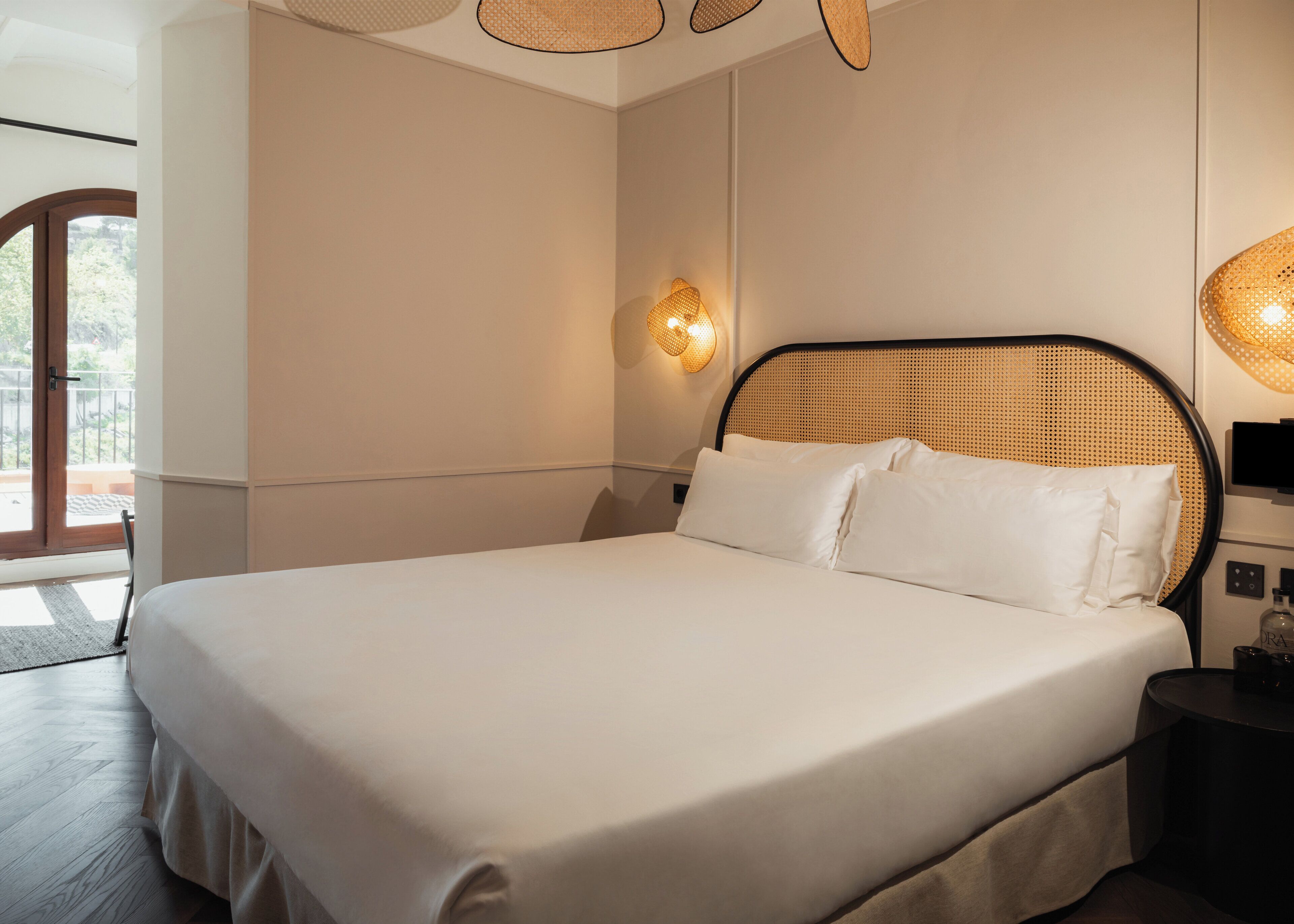 Uma cama grande com lençóis brancos, cabeceira de vime e mesa de cabeceira com uma garrafa.
