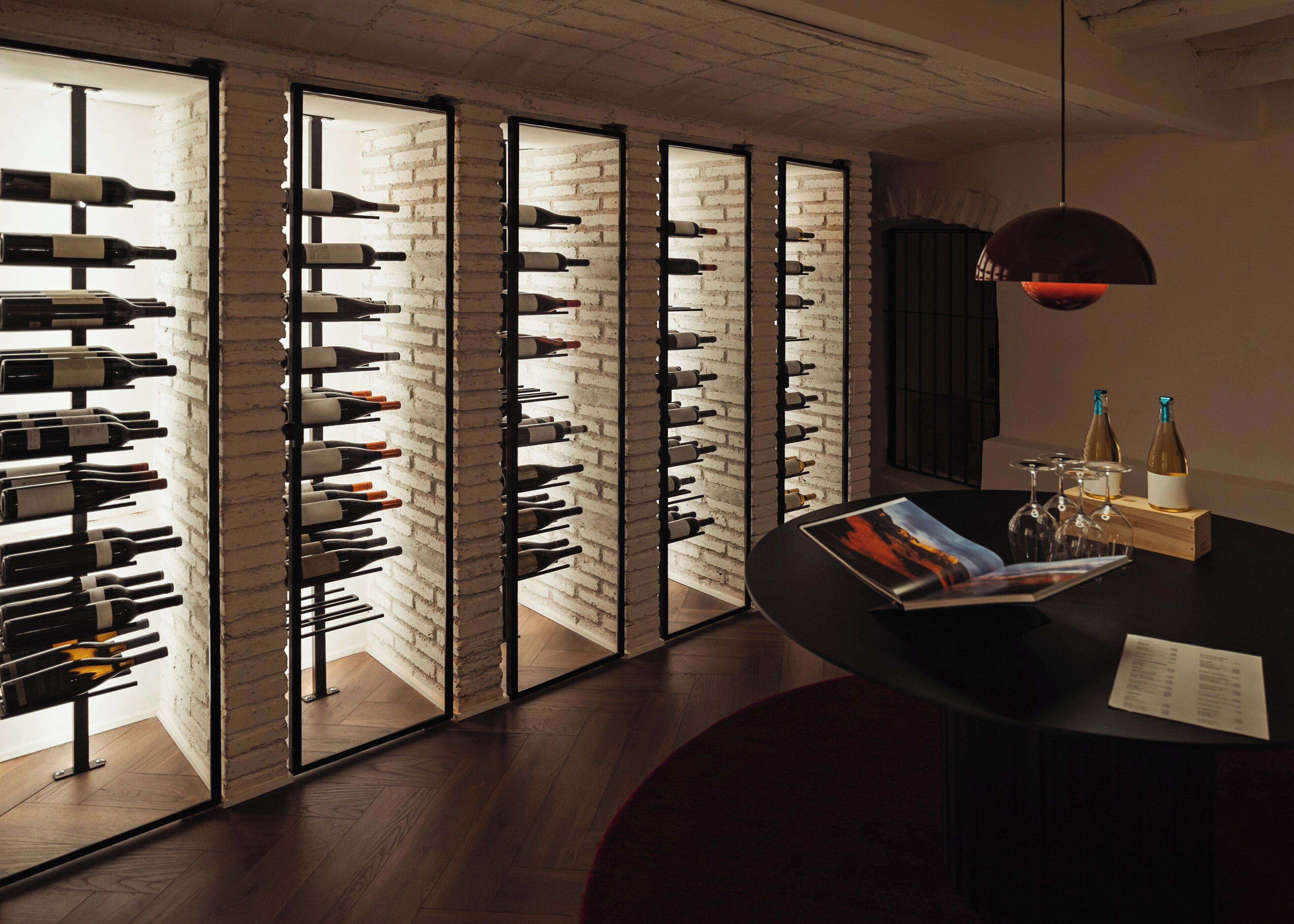A wine cellar with multiple wine racks against a brick wall, a wooden table with wine glasses and bottles, and a hanging light fixture.