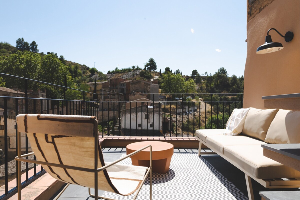 A balcony with a sofa, chair, and round table, overlooking a hillside with buildings.
