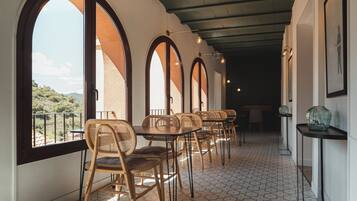 A corridor with arched windows, wicker chairs, and tables, leading to a balcony with a view of the hills.