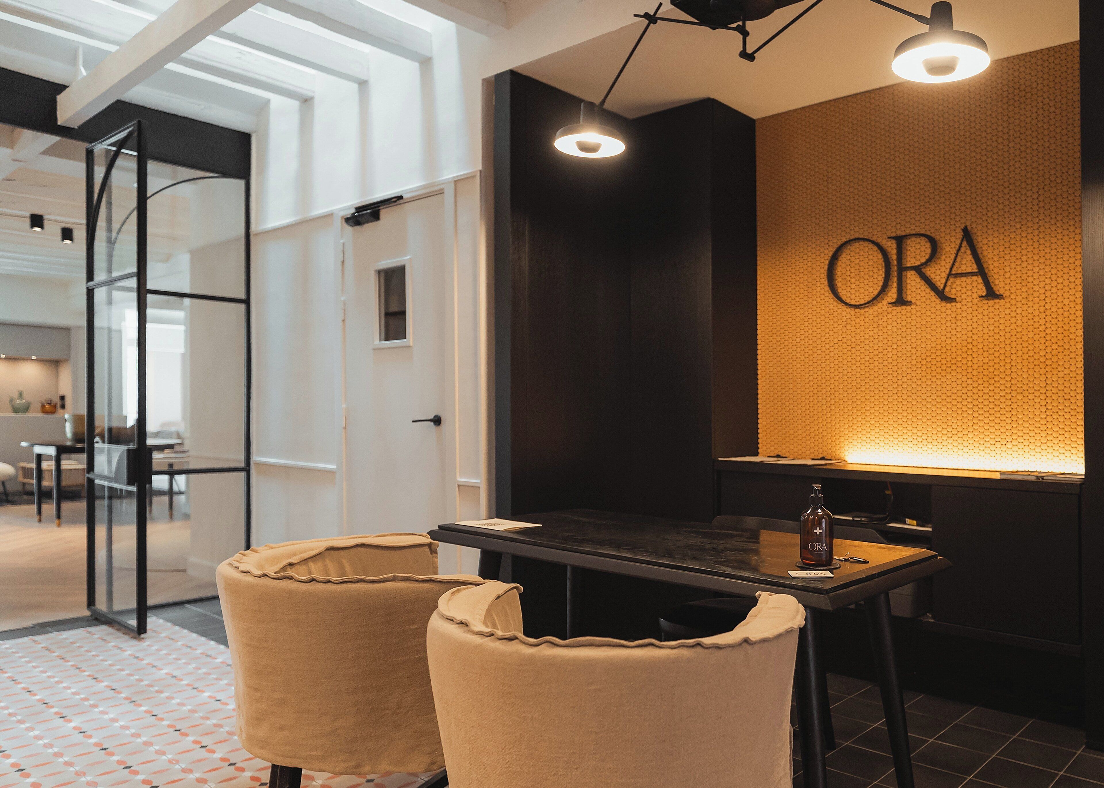A modern interior with a dark wall featuring the word "ORA" in large letters, a table with a bottle, and two upholstered chairs.