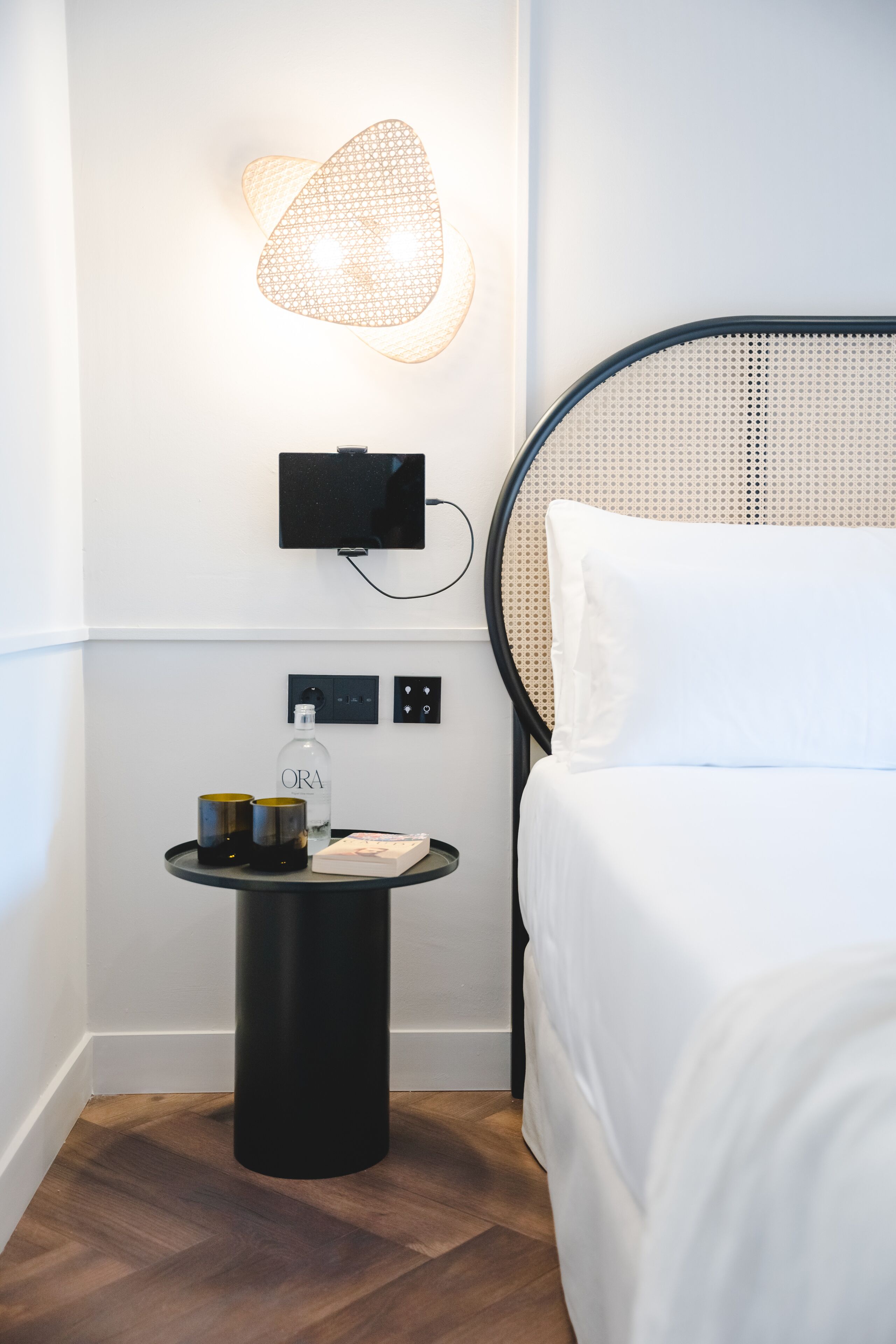 A neatly arranged hotel room with a bed, a bedside table with a bottle and glasses, a wall-mounted TV, and a unique wall light fixture.