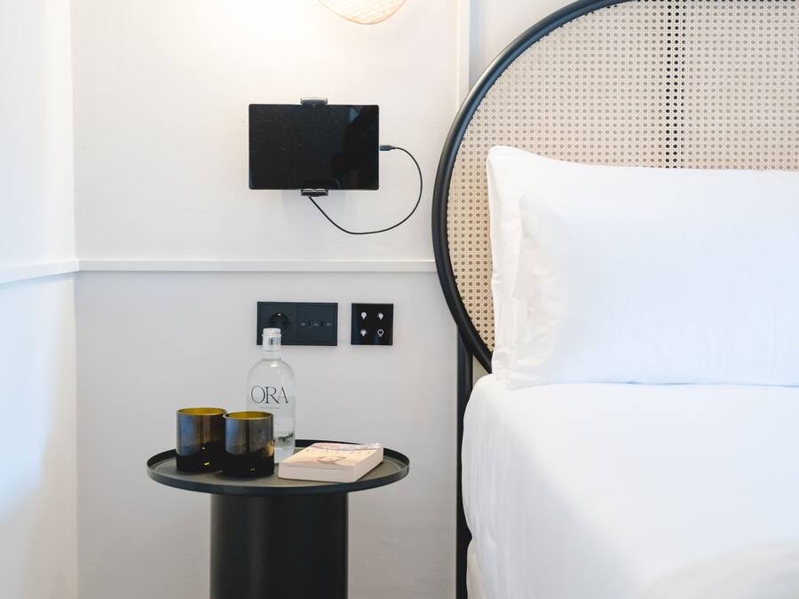 A neatly arranged hotel room with a bed, a bedside table with a bottle and glasses, a wall-mounted TV, and a unique wall light fixture.