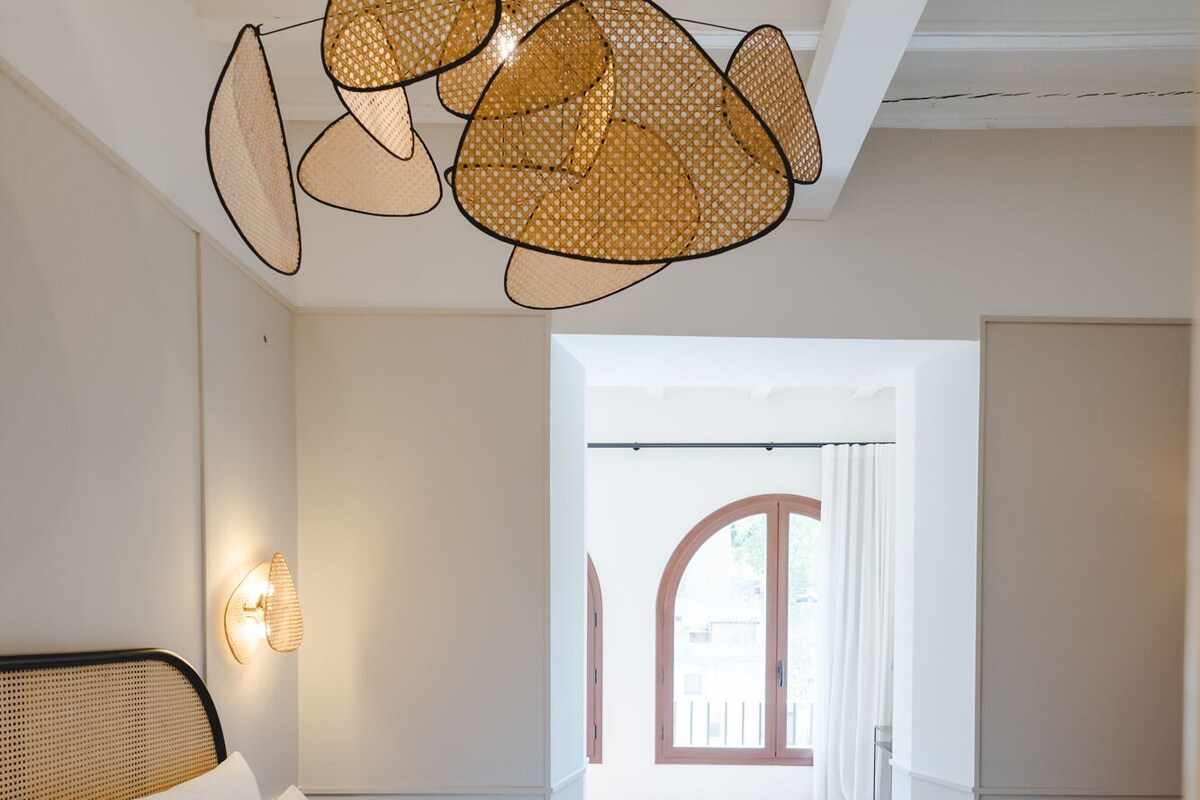 A bedroom with a large bed, a rattan ceiling light, and a view of the outside through a window.