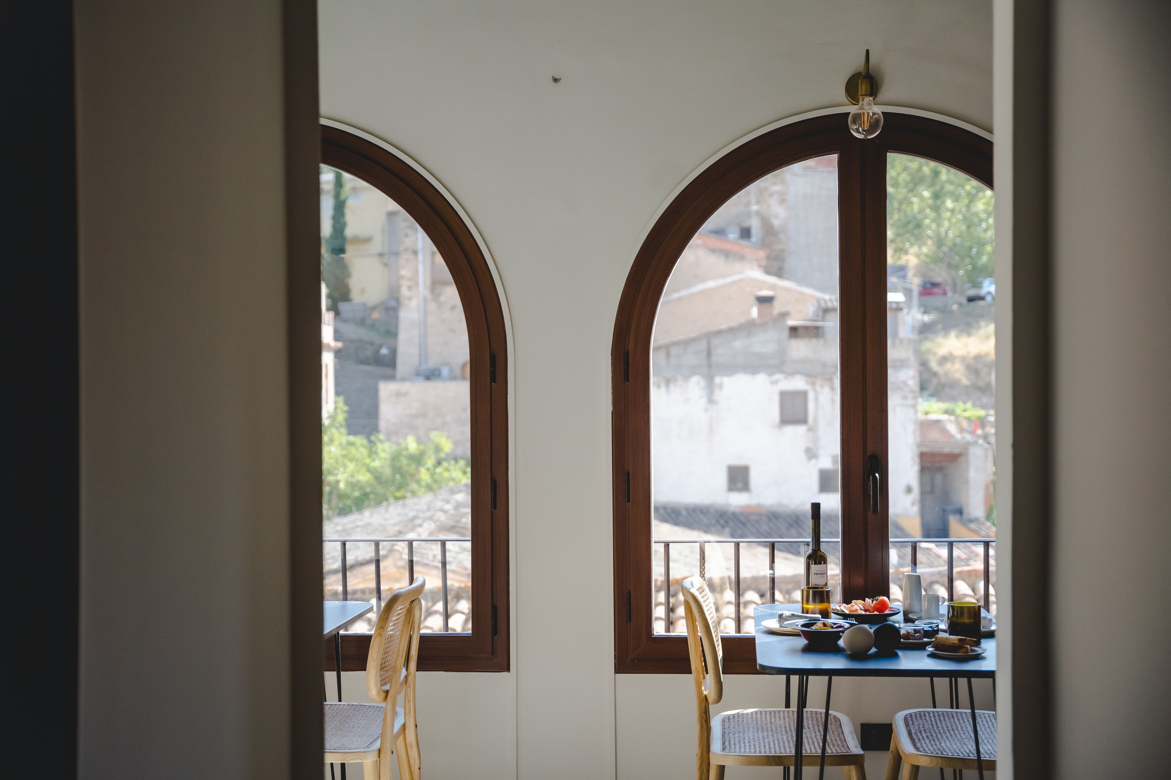 Un comedor con dos ventanas arqueadas, una mesa dispuesta con platos, una botella y sillas.