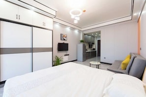 A modern hotel room with a large bed, a sofa, a TV, and a kitchenette.