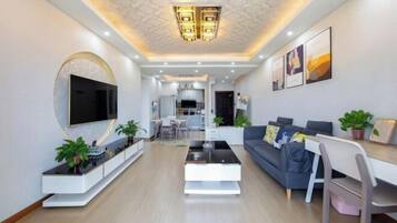 A modern living room with a flat-screen TV, a sofa, a coffee table, and a dining area with a white table and chairs.