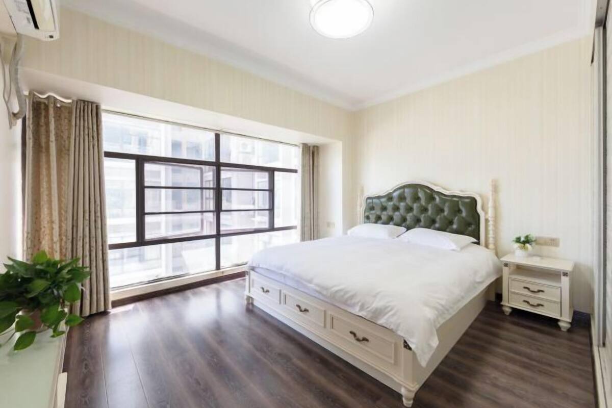 A bedroom with a large window, a bed with a tufted headboard, a bedside table, and a nightstand.