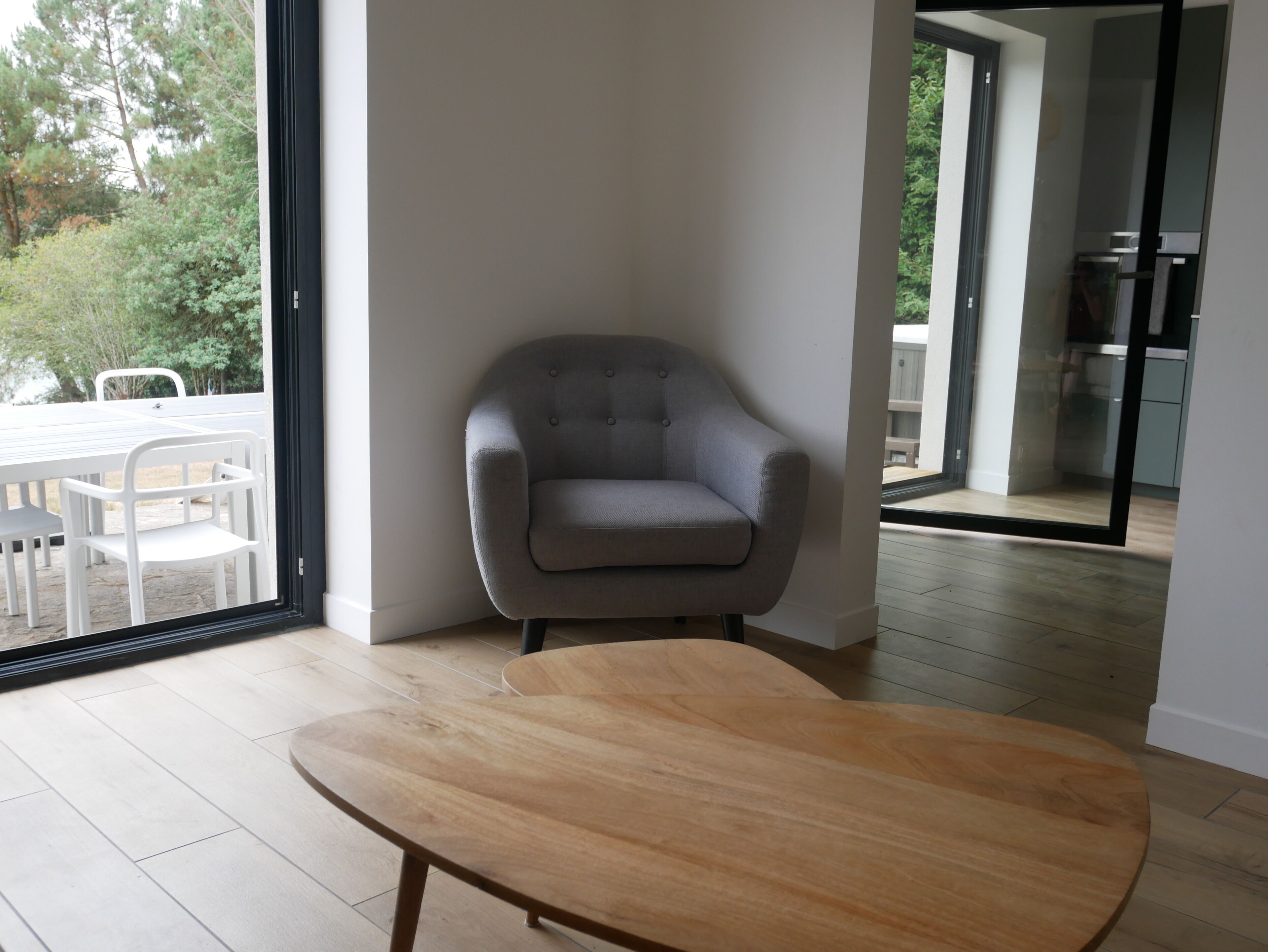 Un salon moderne avec un fauteuil gris, une table basse en bois et une porte vitrée donnant sur un balcon aménagé avec du mobilier d’extérieur.