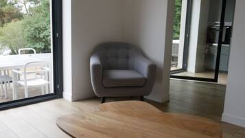Un salon moderne avec un fauteuil gris, une table basse en bois et une porte vitrée donnant sur un balcon aménagé avec du mobilier d’extérieur.
