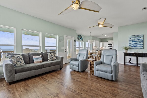 A spacious living room with a grey sofa, two armchairs, and a coffee table. There are large windows with a view of the beach, a ceiling fan, and a kitchen in the background.