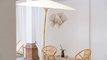 A room with a large umbrella table lamp, two wicker chairs, a small table with a plant, and a decorative wall piece.