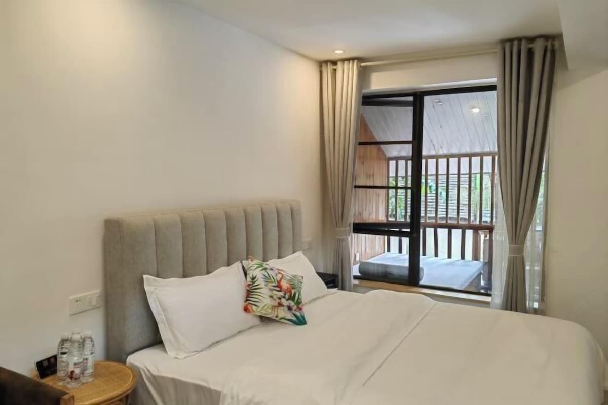 A hotel room with a bed, a nightstand, a window with curtains, and a view of a balcony.