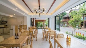 Ein Restaurant mit Holztischen und -stühlen, einer Kristallleuchtre und großen Fenstern mit Blick auf die Straße und die Grünflächen davor.