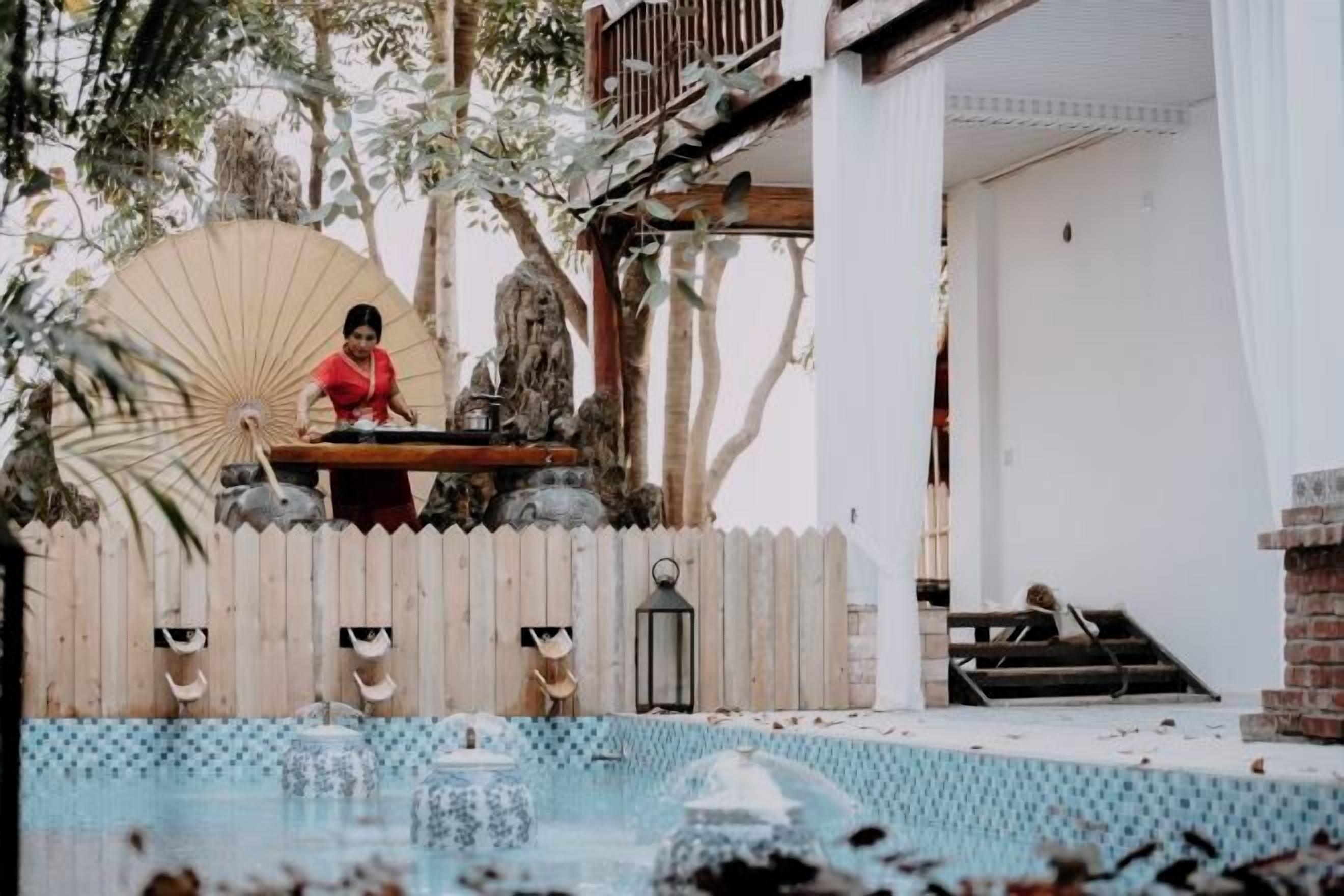 Área de piscina com deck de madeira, uma pessoa de camisa vermelha e um guarda-sol grande.