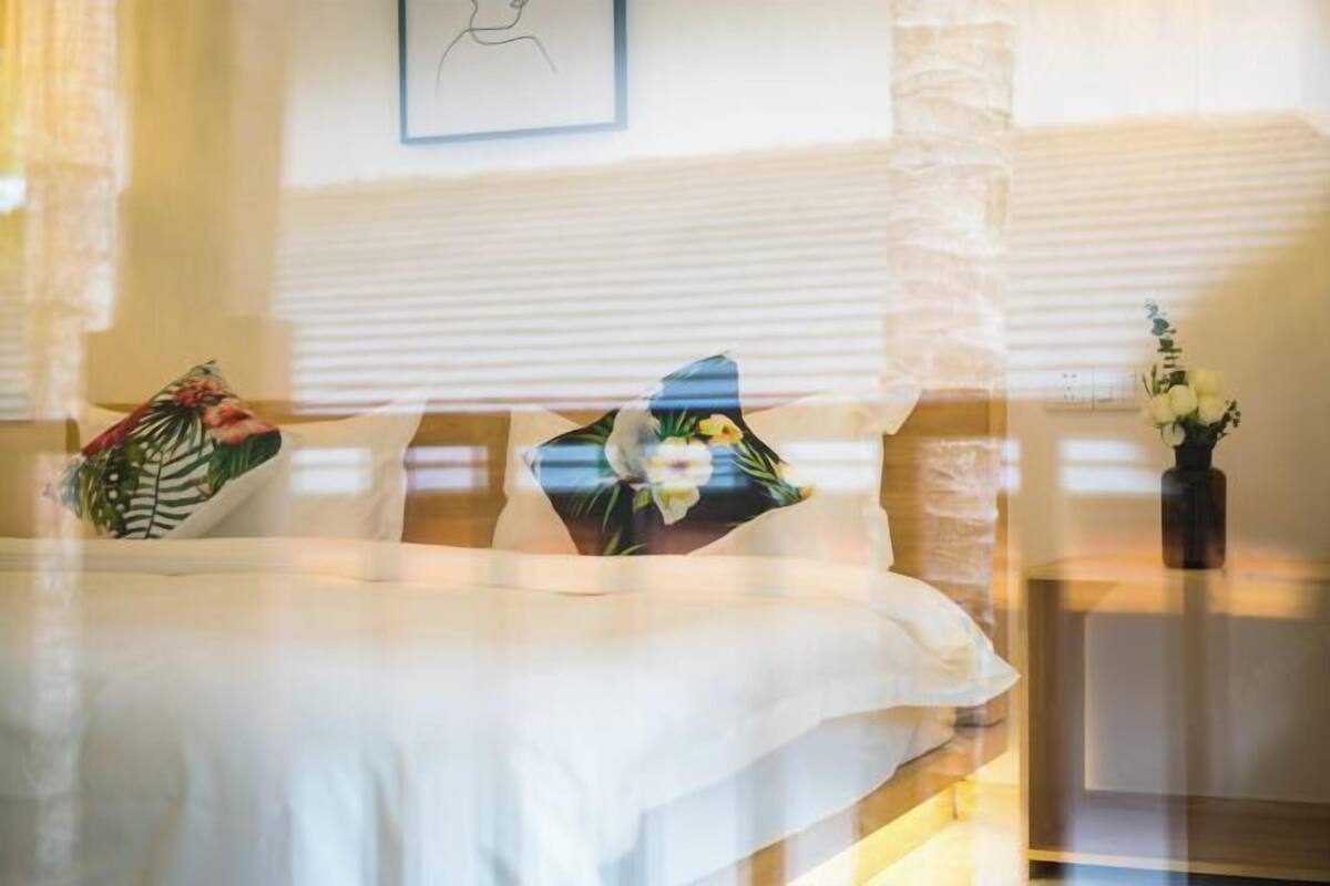 Een hotelkamer met een bed, kussens met bloemenmotieven, een ingelijst foto aan de muur en een vaas met bloemen op een nachtkastje.