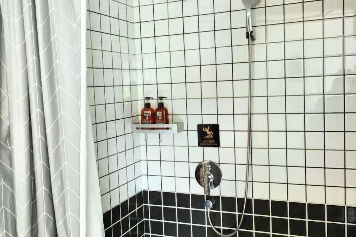 A bathroom with white and black tiled walls, a shower area, and a shelf with toiletries.