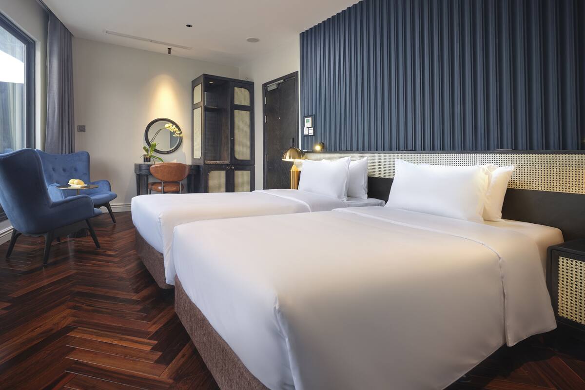 A hotel room with two beds, a blue armchair, a small table with a lamp, and a large window.
