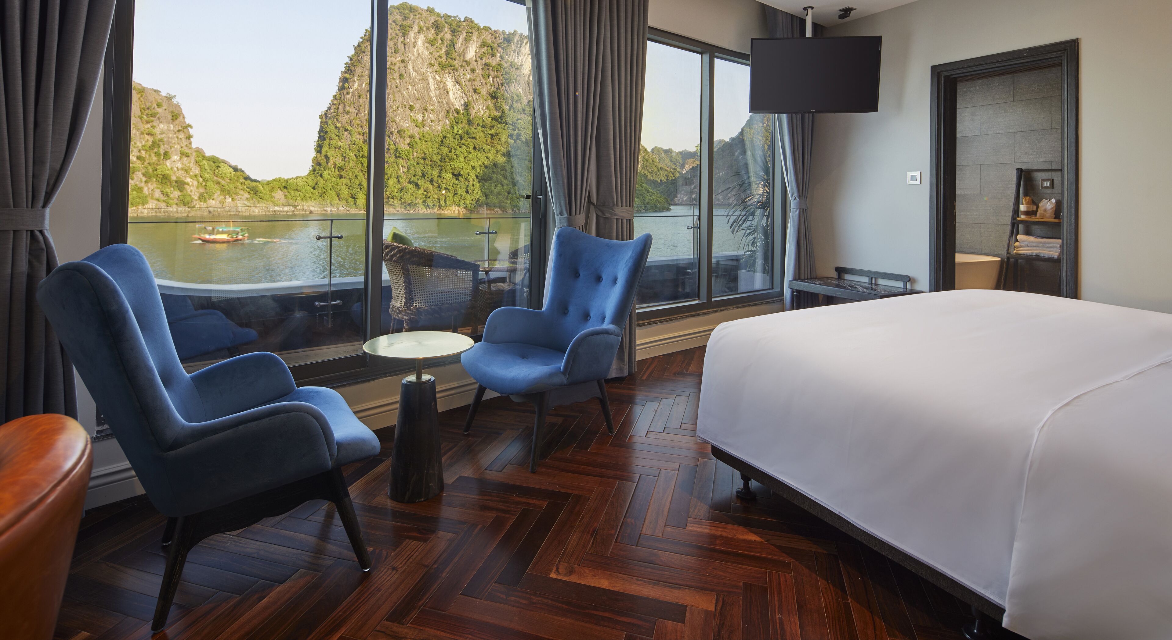 A hotel room with a large bed, two blue armchairs, a small round table, and a view of a lake and mountains through large windows.