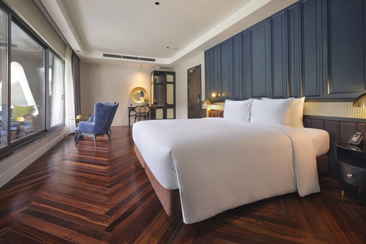 A hotel room with a large bed, a desk with a chair, a mirror, and a large window.