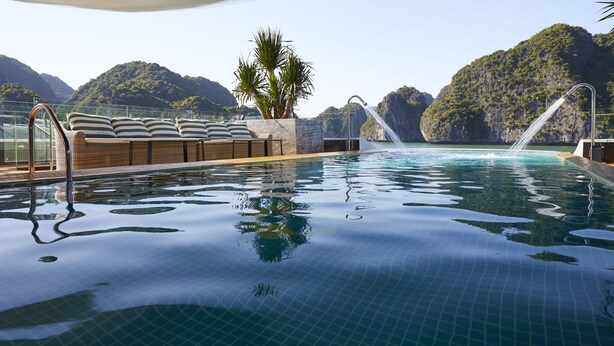 A pool with a waterfall feature, surrounded by lounge chairs and a view of mountains. - Elite of the Seas (Tuan Chau)