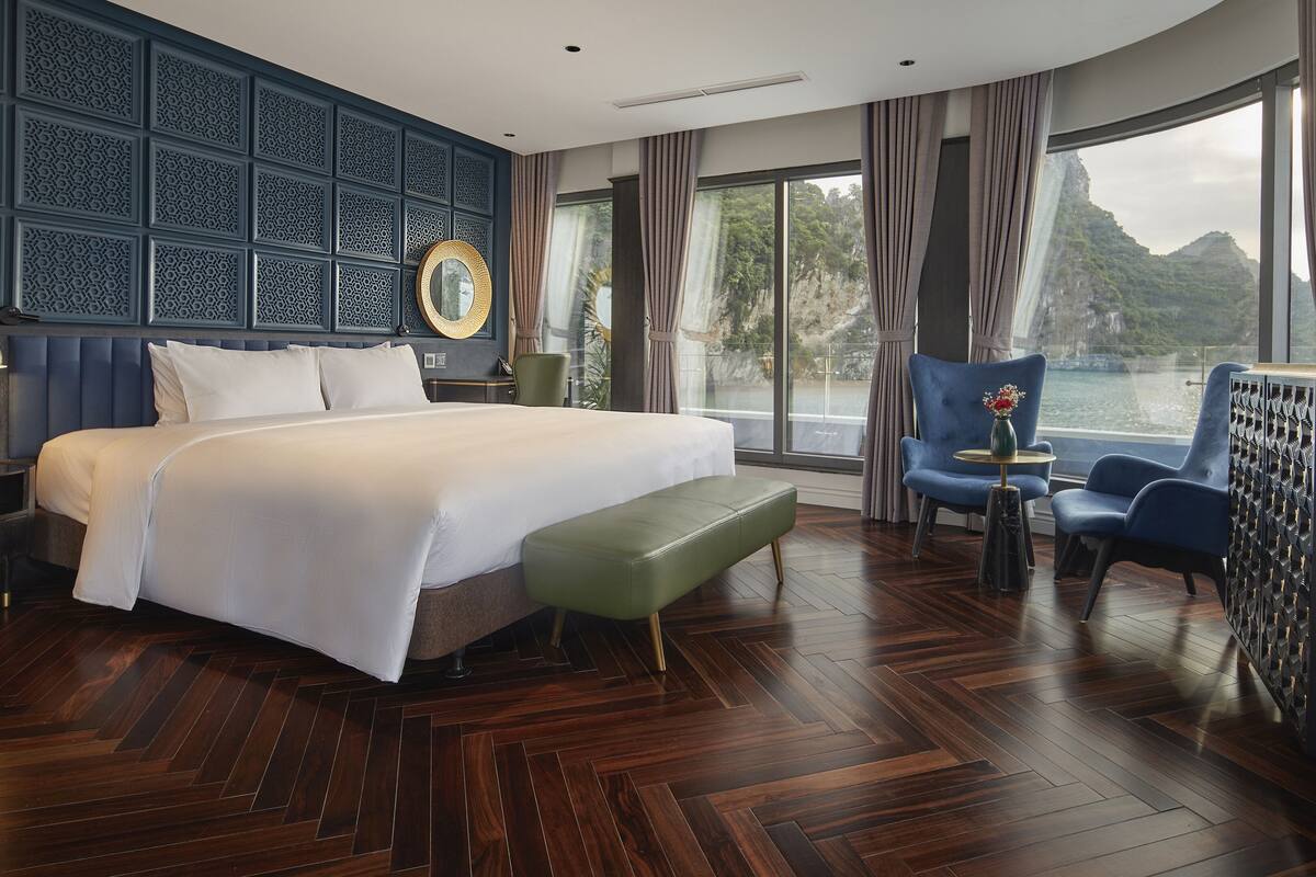 A modern hotel room with a large bed, a green bench, a blue armchair, and a view of a mountainous landscape through large windows.