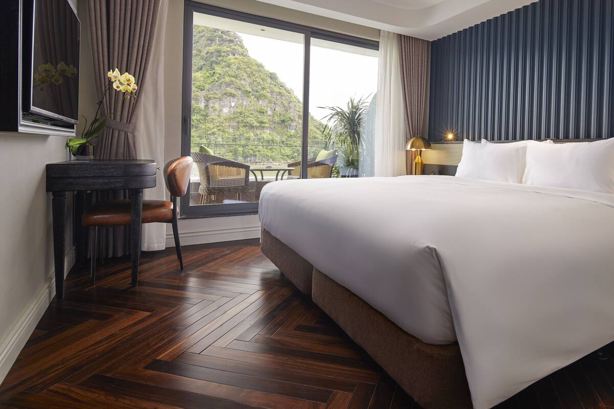 A hotel room with a large bed, a desk with a chair, a view of a mountain, and a balcony with wicker furniture.