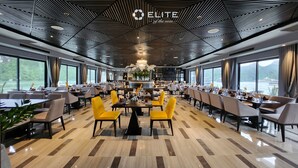A modern dining area with a geometric ceiling design, large windows offering views of the sea, and a central chandelier. - Elite of the Seas (Tuan Chau)