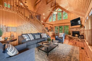 A spacious living room with a large sofa, a coffee table, and a fireplace. A staircase leads to an upper level, and there is a TV mounted on the wall.