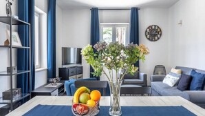 Una sala de estar moderna con una mesa de comedor, una cesta de frutas, un florero con flores, un sofá, un televisor y un reloj en la pared.
