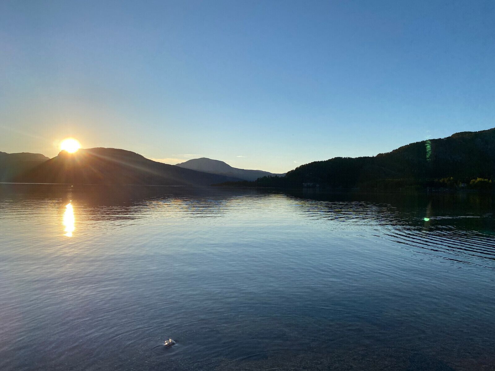 Un lago tranquilo con montañas al fondo y el sol poniéndose en el horizonte.