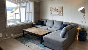 En moderne stue med en grå hjørnesofa, et træsofabord og et stort vindue med udsigt til et bjerg.