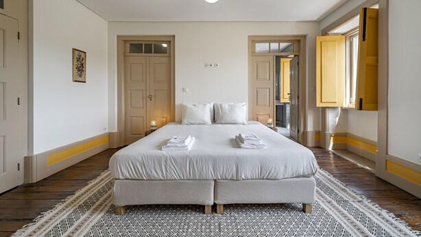 Uma cama grande com roupa de cama branca, um tapete estampado e um candeeiro suspenso.