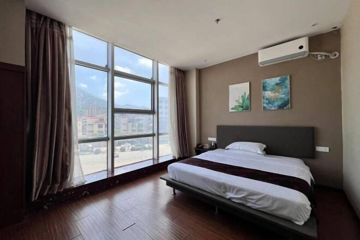 A hotel room with a large window, a bed, a nightstand, and a wall-mounted air conditioner.