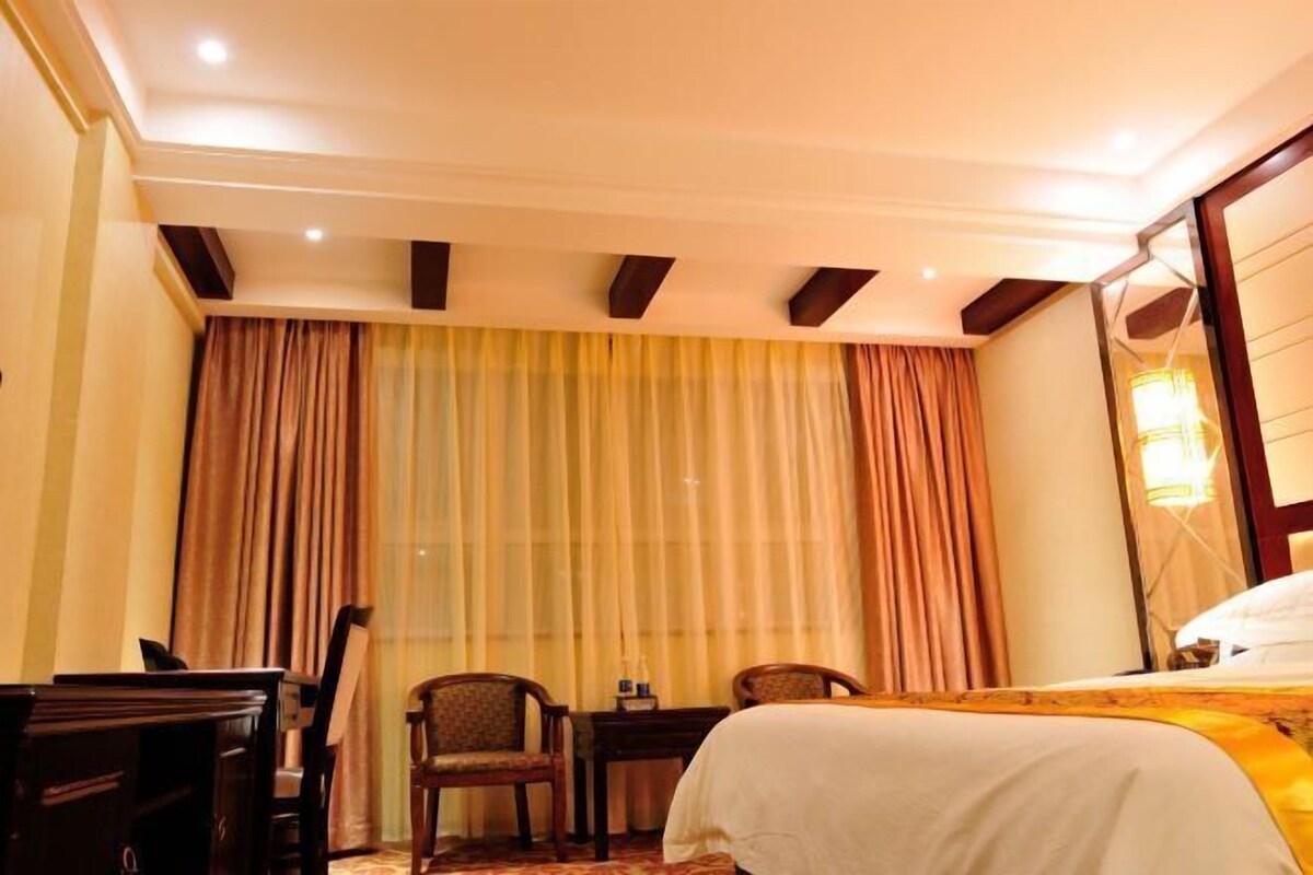 A hotel room with a bed, a desk, two chairs, a window with curtains, and a lamp.