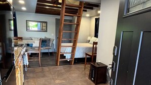 Une cuisine compacte avec un sol carrelé en pierre, une échelle en bois, un lit recouvert d’une courtepointe à motifs et une petite table avec une lampe.
