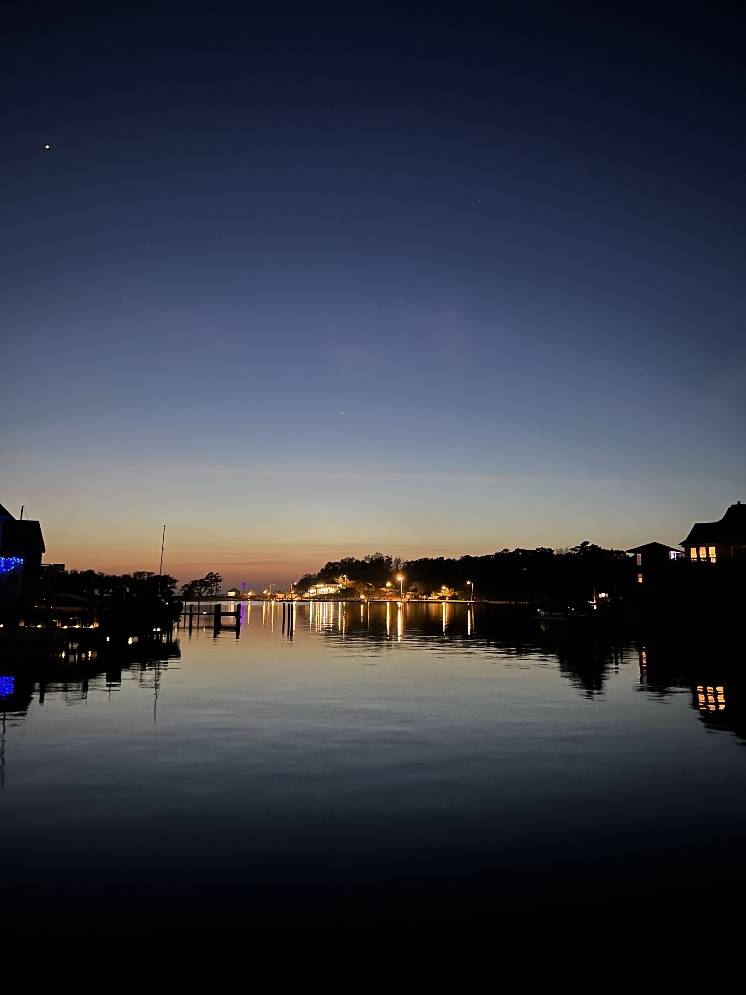 Un paysage paisible au bord de l’eau, au crépuscule, avec des bâtiments illuminés et un ciel dégagé.