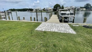 A dock leading to a boat, surrounded by a grassy area and a calm lake with houses in the background. - Waterfront Harbor Home, Sunset Views, Fireplace, Private dock and Boat Slip (Kill Devil Hills)
