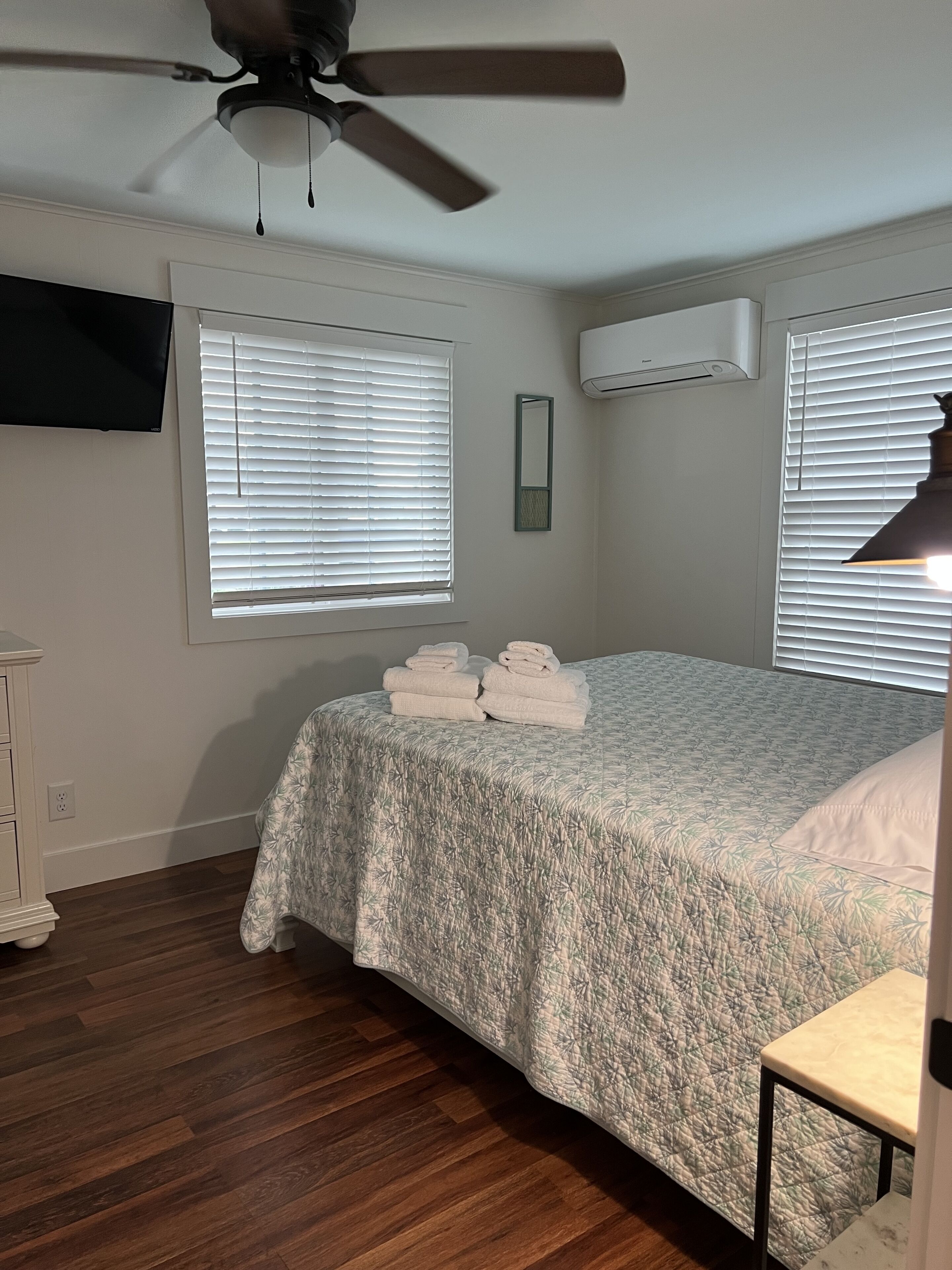 A bedroom with a bed, a ceiling fan, a TV mounted on the wall, and a window with blinds.