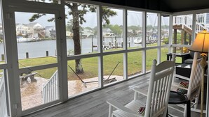 A covered porch with a view of a lake and houses. There are white chairs and a table with a lamp. - Waterfront Harbor Home, Sunset Views, Fireplace, Private dock and Boat Slip (Kill Devil Hills)