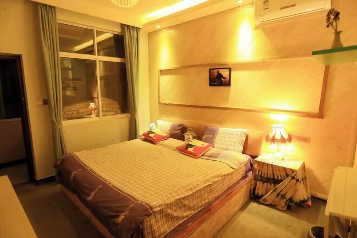 A hotel room with a large bed, a bedside table with a lamp, a window with curtains, and a wall-mounted picture.