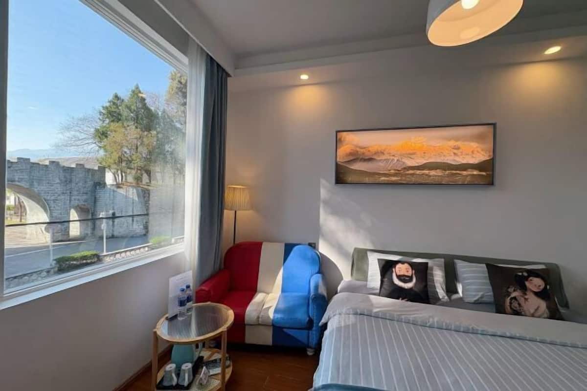 A hotel room with a bed, a sofa, a small table, a lamp, a painting, and a view of a stone bridge and trees.