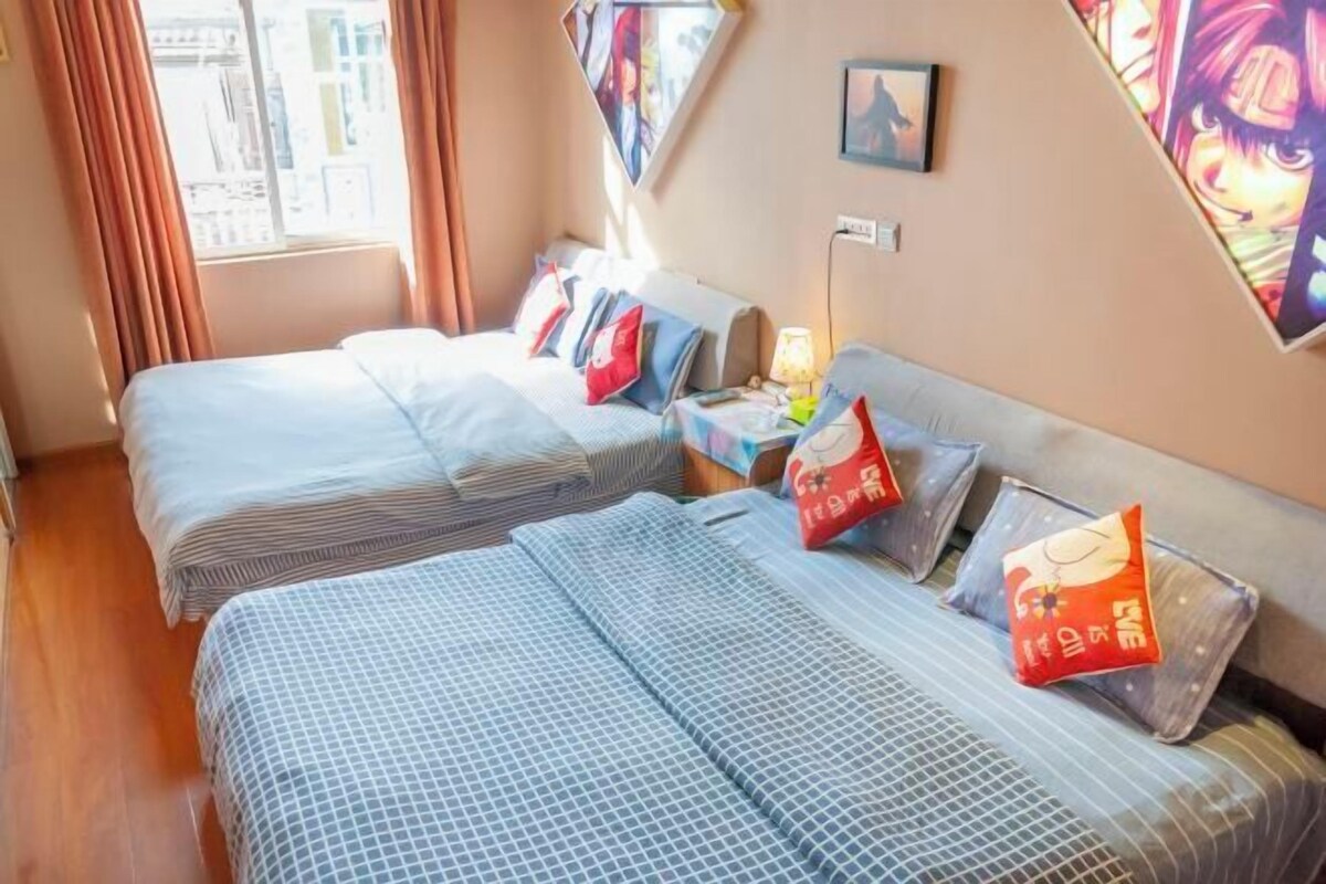 A hotel room with two beds, each with patterned bedding and decorative pillows. There is a nightstand with a lamp and a small table with a phone and a book. The room has a window with curtains and a view of buildings outside.