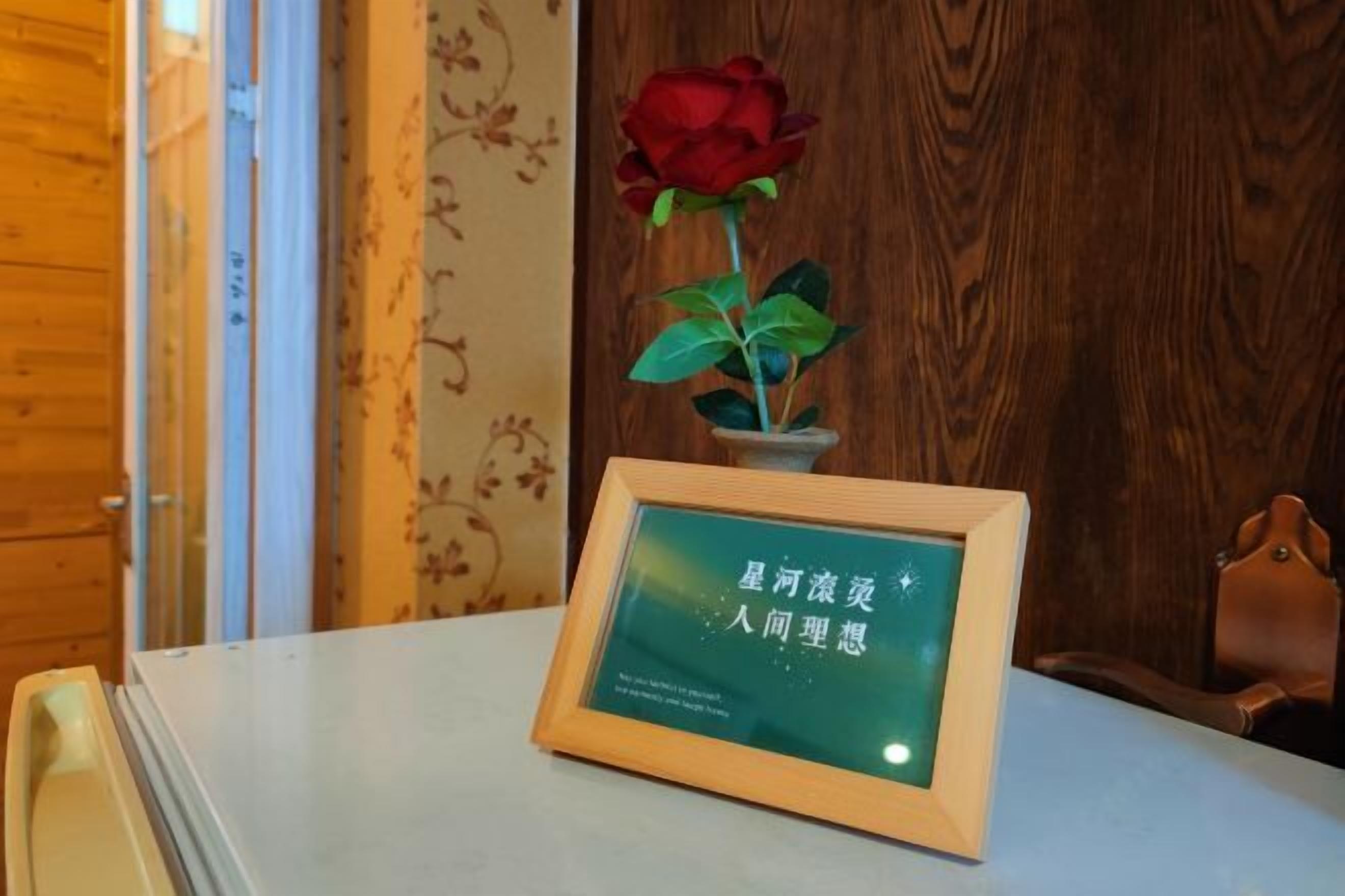Un letrero enmarcado con caracteres chinos sobre una mesa, una rosa roja en maceta y una pared de madera al fondo.