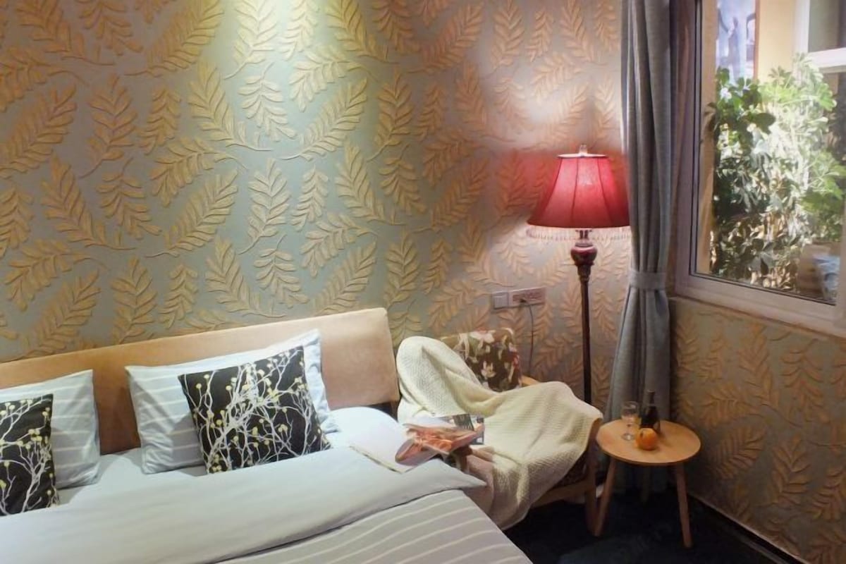 A hotel room with a bed, a bedside table with a lamp, and a window with curtains.