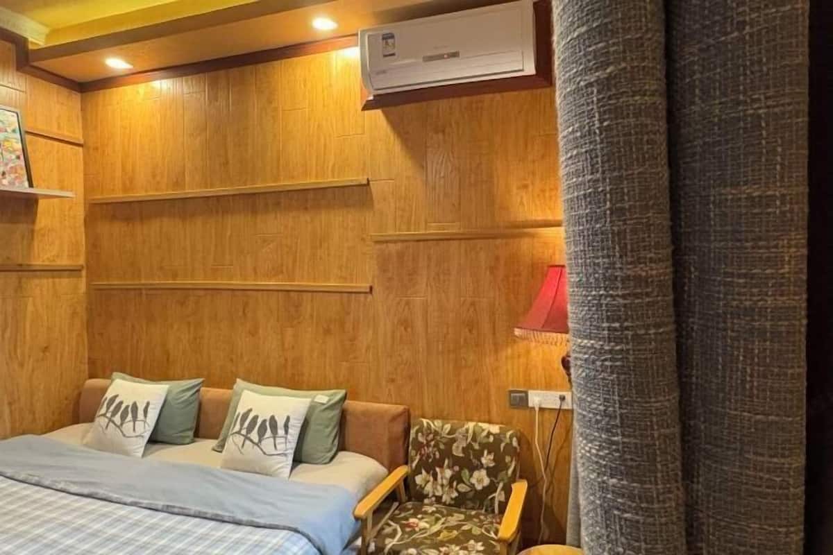 A room with wooden panel walls, a bed with a blue bedspread and pillows, a floral armchair, and a red lamp.