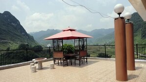 A terrace with a table and chairs under a red umbrella, a stone planter, and a view of mountains.