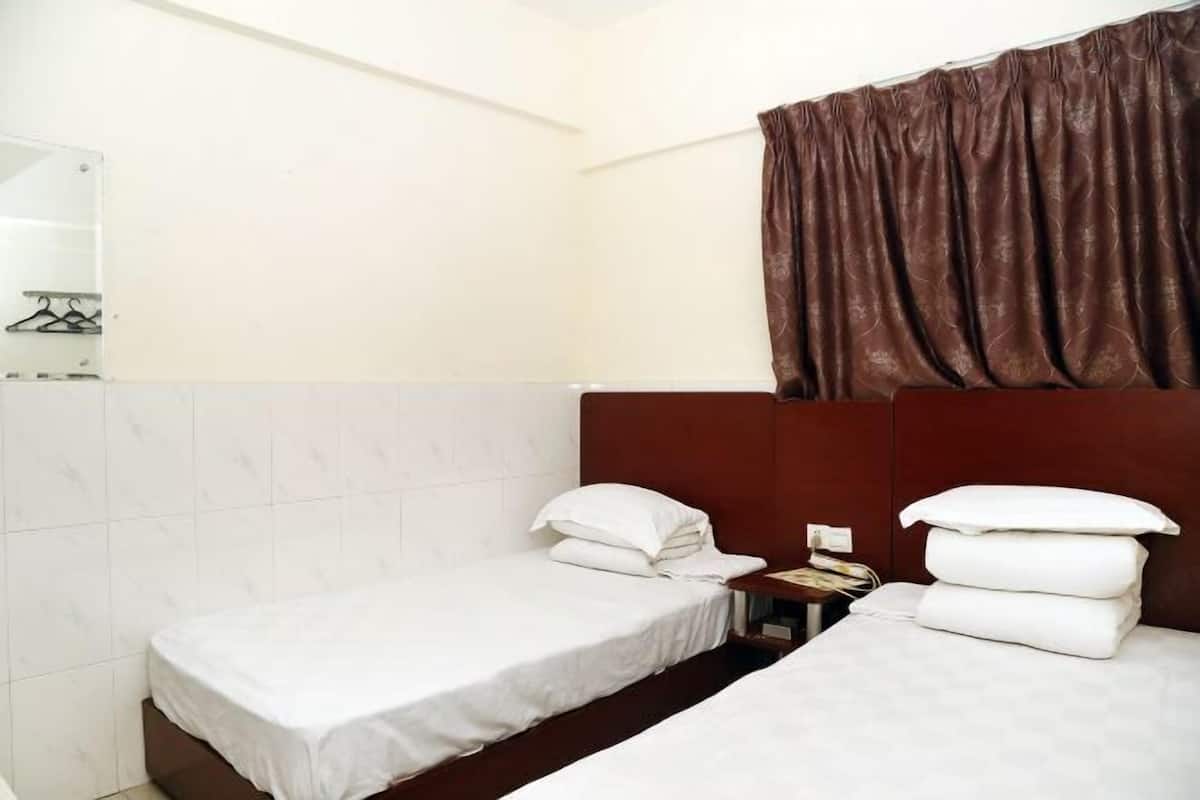A hotel room with two single beds, a wooden headboard, a small bedside table with a lamp, and a brown curtain.