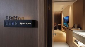 Une chambre d’hôtel avec une porte en bois, un écran numérique affichant le numéro de chambre 890808, et une salle de bain moderne équipée d’une télévision et d’un grand miroir.